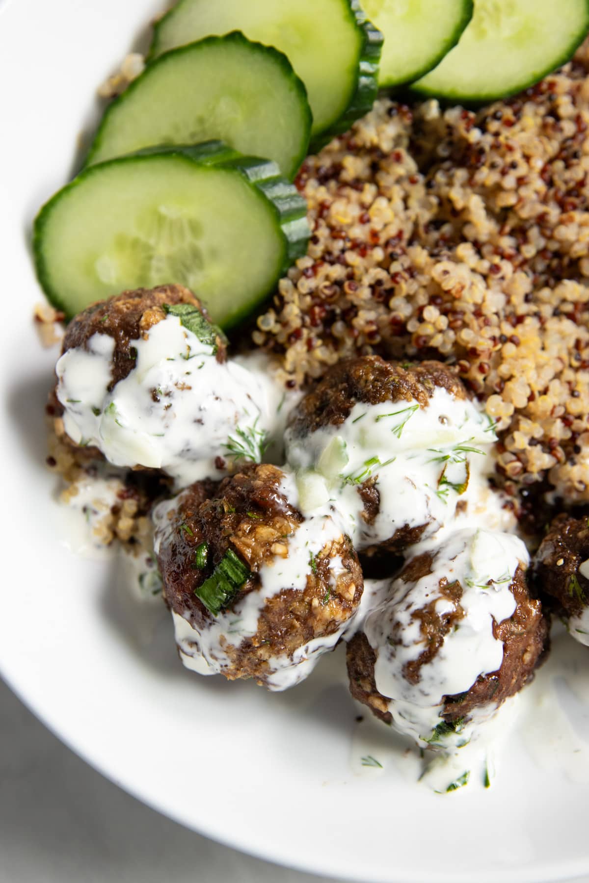 Focused on a handful of beef meatballs drizzled with a creamy dill sauce and sitting next to cooked tri-color quinoa and cucumber slices.