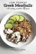 A shallow bowl filled with cooked quinoa, low FODMAP Greek meatballs topped with a creamy dill sauce, and a few cucumber slices on the side. In the white space above, black text reads "Low FODMAP Greek Meatballs with creamy cucumber-dill sauce."