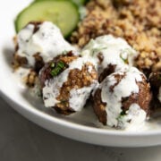 Focus on a bowl of Low FODMAP Greek Meatballs drizzled with Creamy Cucumber Dill Sauce. Cooked quinoa and cucumber slices are blurred in the background.