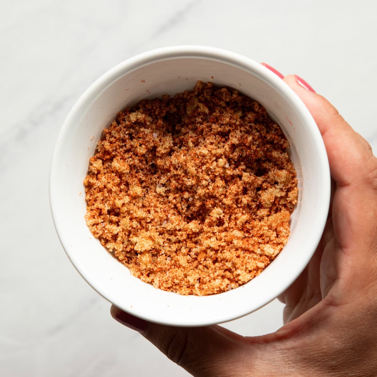 A hand holding a small white dish containing a rub made from brown sugar, smoked paprika, and salt.