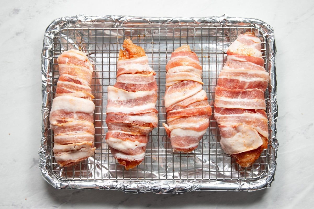 Four chicken breasts wrapped in bacon on a wire rack sitting in a sheet pan lined with aluminum foil.