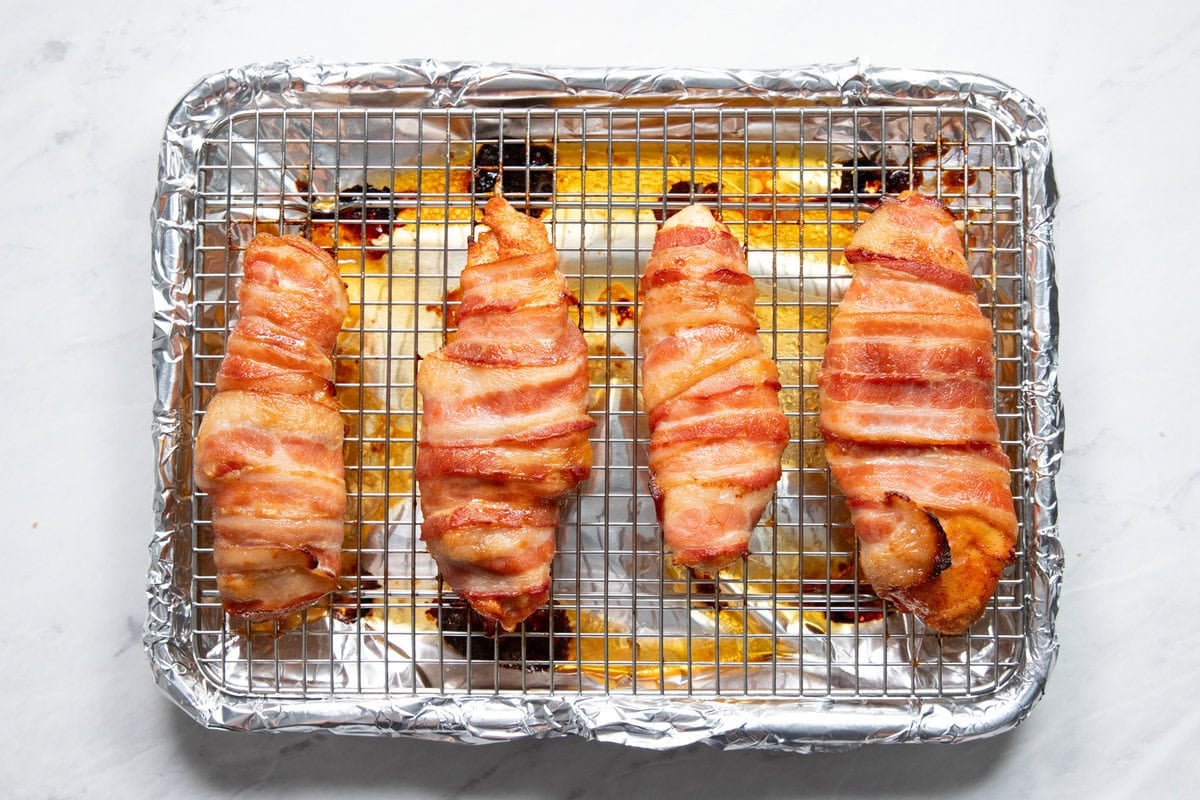 Four baked bacon wrapped chicken breasts sitting on a wire rack in a sheet pan lined with aluminum foil.