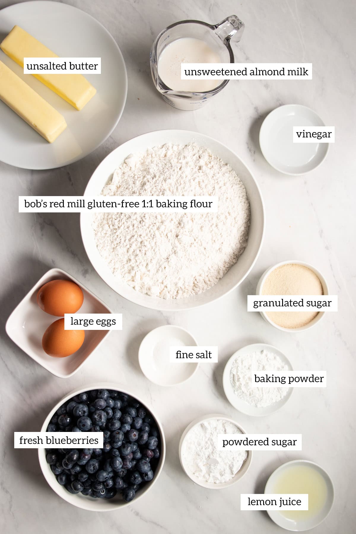 Ingredients needed for low FODMAP blueberry scones are prepared and measured out into individual dishes on a white marble countertop.