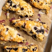 Five blueberry scones drizzled with a lemon glaze are sitting on a parchment lined baking sheet.