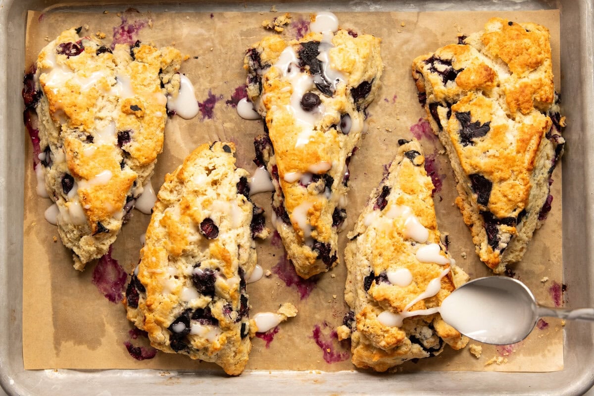 Drizzling lemon glaze with a spoon over five baked low FODMAP blueberry scones on a parchment lined baking sheet. 