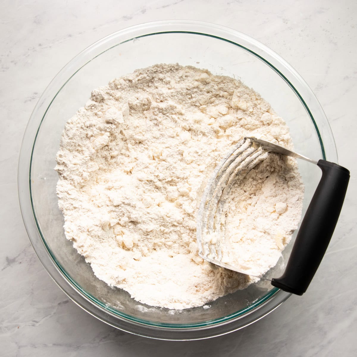 A large mixing bowl containing pea-sized pieces of cold butter cut into a flour mixture using a pastry cutter that's resting inside the bowl.