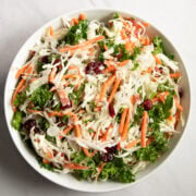 Looking down into a big white bowl filled with a salad made with shredded green cabbage, kale, carrots, dried cranberries, green onion tops, and sunflower seeds.