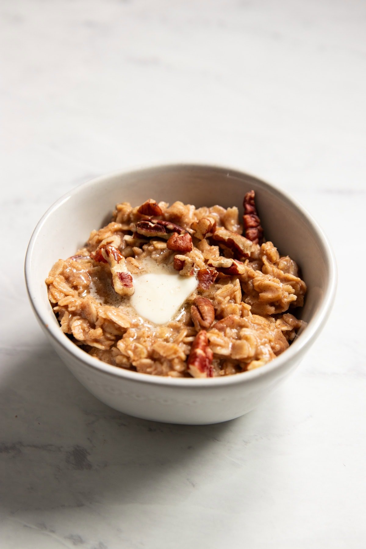 Looking into a bowl of warm oatmeal. It's topped with a dab of melting butter and crunchy pecans.