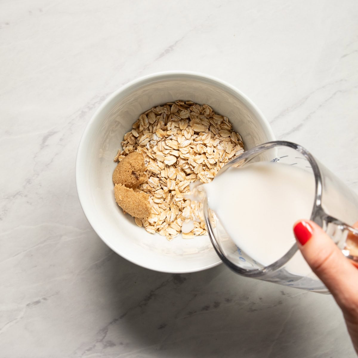 Pouring almond milk into a microwavable-safe bowl containing rolled oats, brown sugar, and pinch of salt.