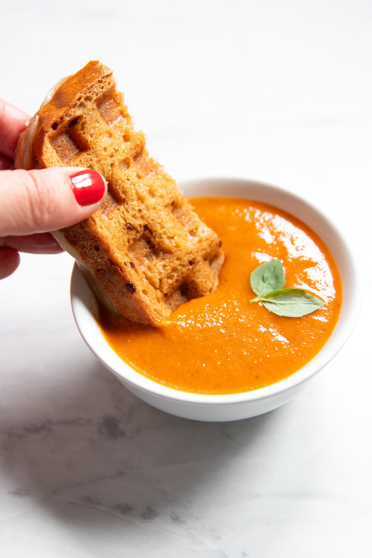 A piece of waffle-maker grilled cheese getting dipped into a bowl of low-FODMAP tomato soup.