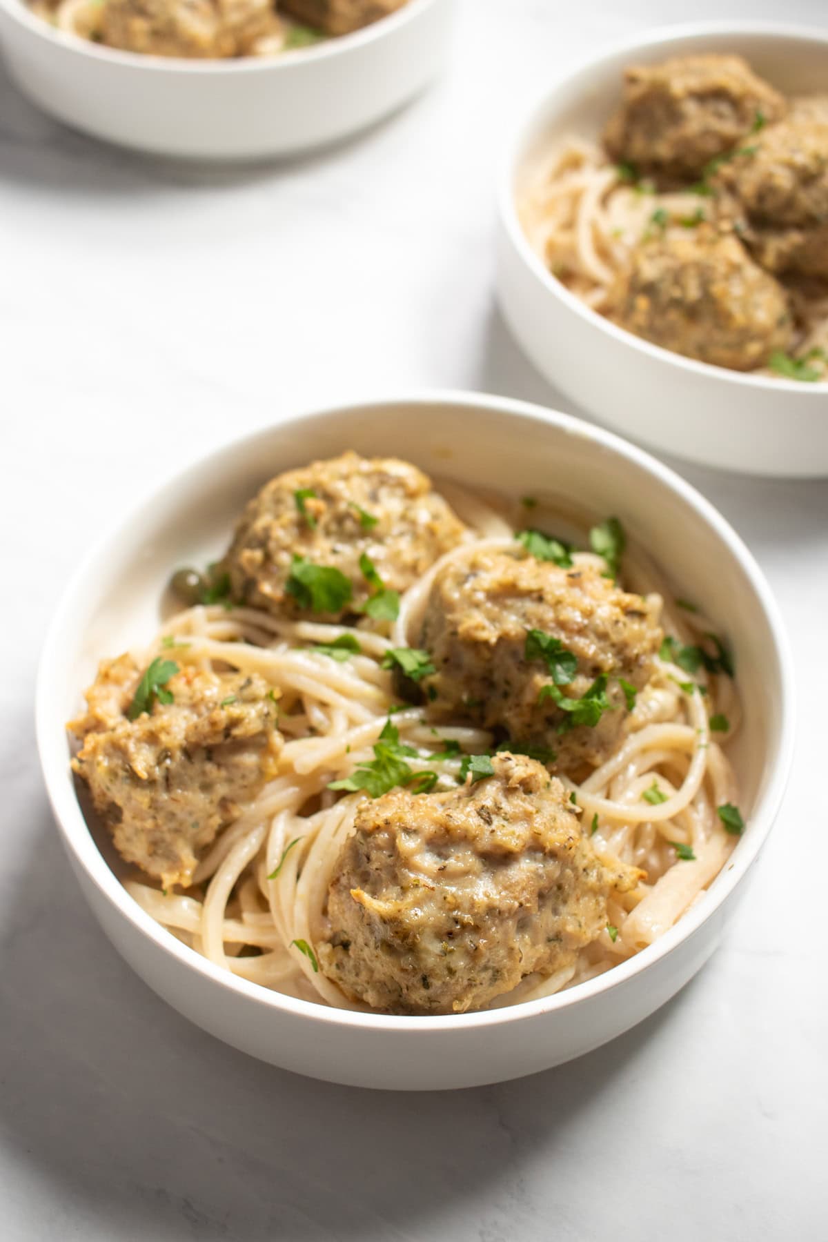 Three bowls of low FODMAP chicken piccata meatballs and spaghetti are staggered on a white marble countertop.