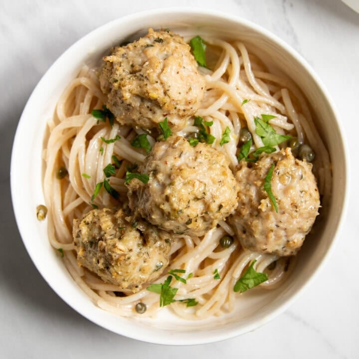 Looking down at a shallow white bowl filled with gluten-free spaghetti tossed in a lemon-caper piccata sauce and topped with four chicken meatballs and fresh parsley.