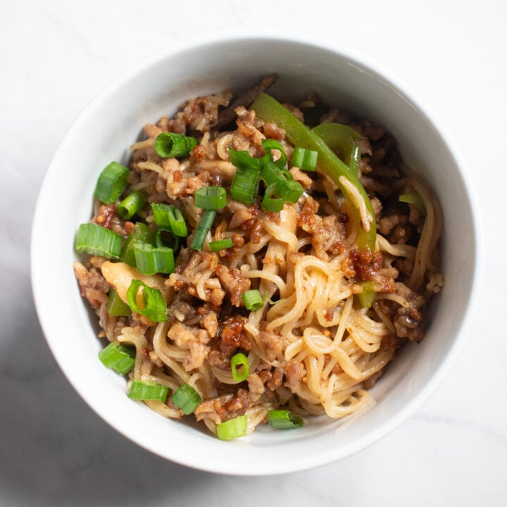 Looking down into a bowl filled with brown rice ramen tossed in an Asian-inspired sauce with ground pork, sauteed green bell peppers, oyster mushrooms and sliced green onion tops.