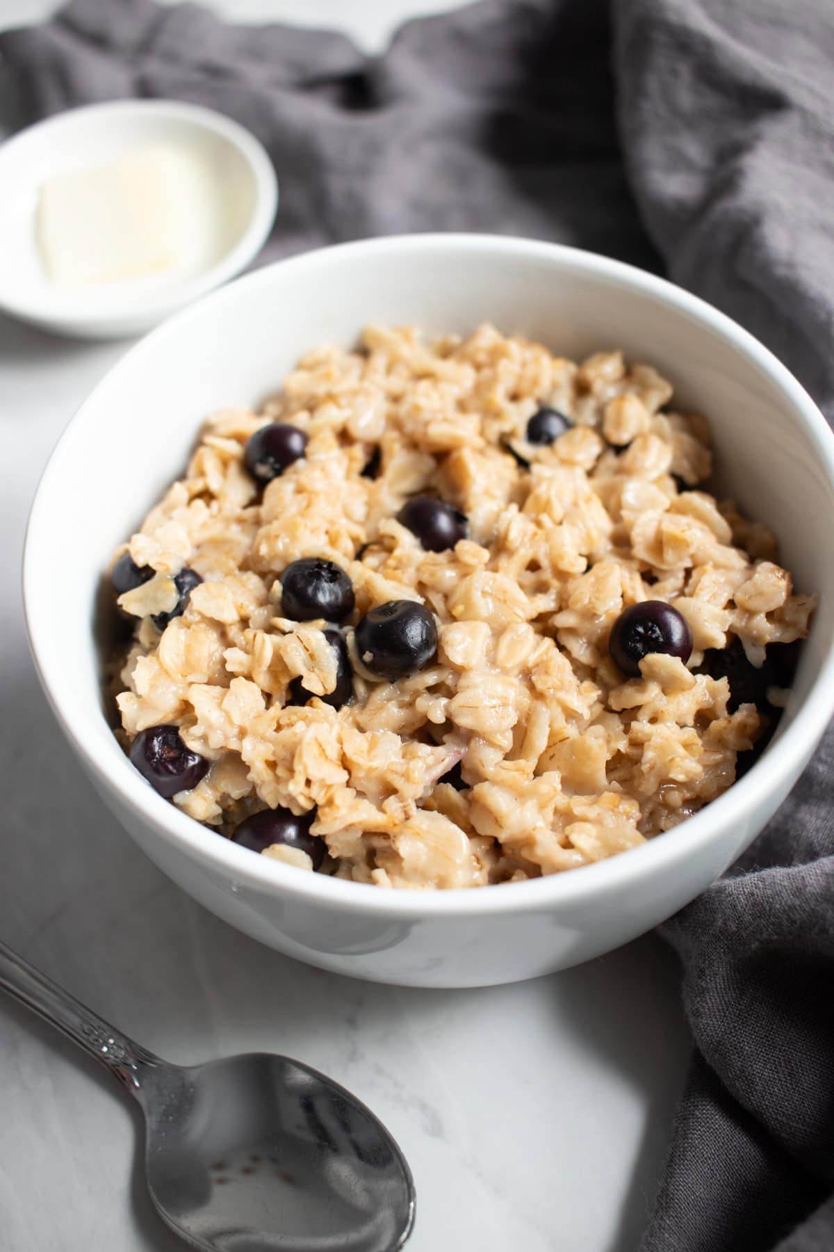 A bowl of blueberry-vanilla flavored oatmeal sits on a white marble counter surrounded by a spoon, a small dish of butter, and a dark gray linen napkin.