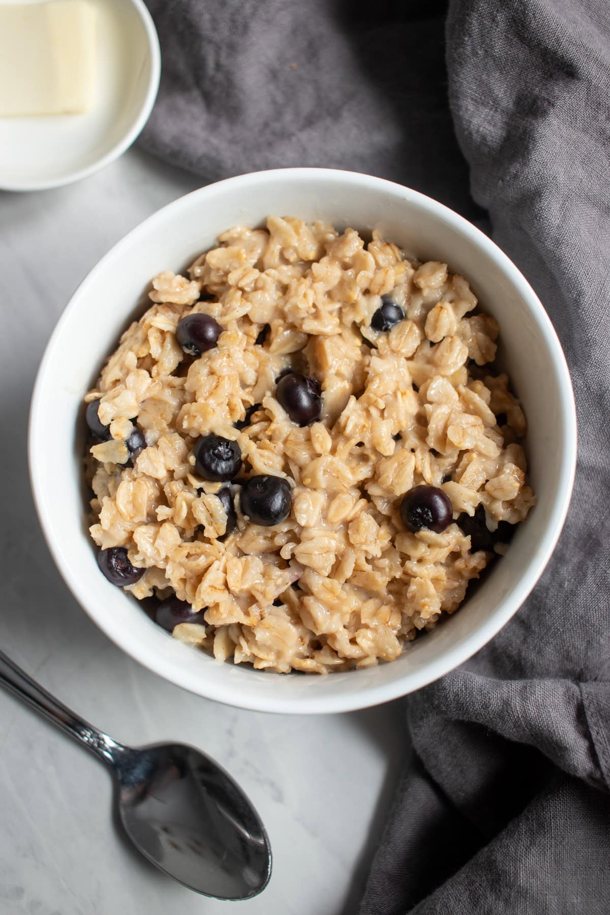 Looking down into a bowl of cooked oatmeal dotted with blueberries. Surrounding the bowl is a dark gray linen napkin, an antique looking spoon, and small dish containing a thin slice of butter.