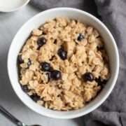 Looking down into a bowl of cooked oatmeal dotted with blueberries. The bowl is surrounded by a dark gray napkin, an antique-looking spoon, and a small dish.