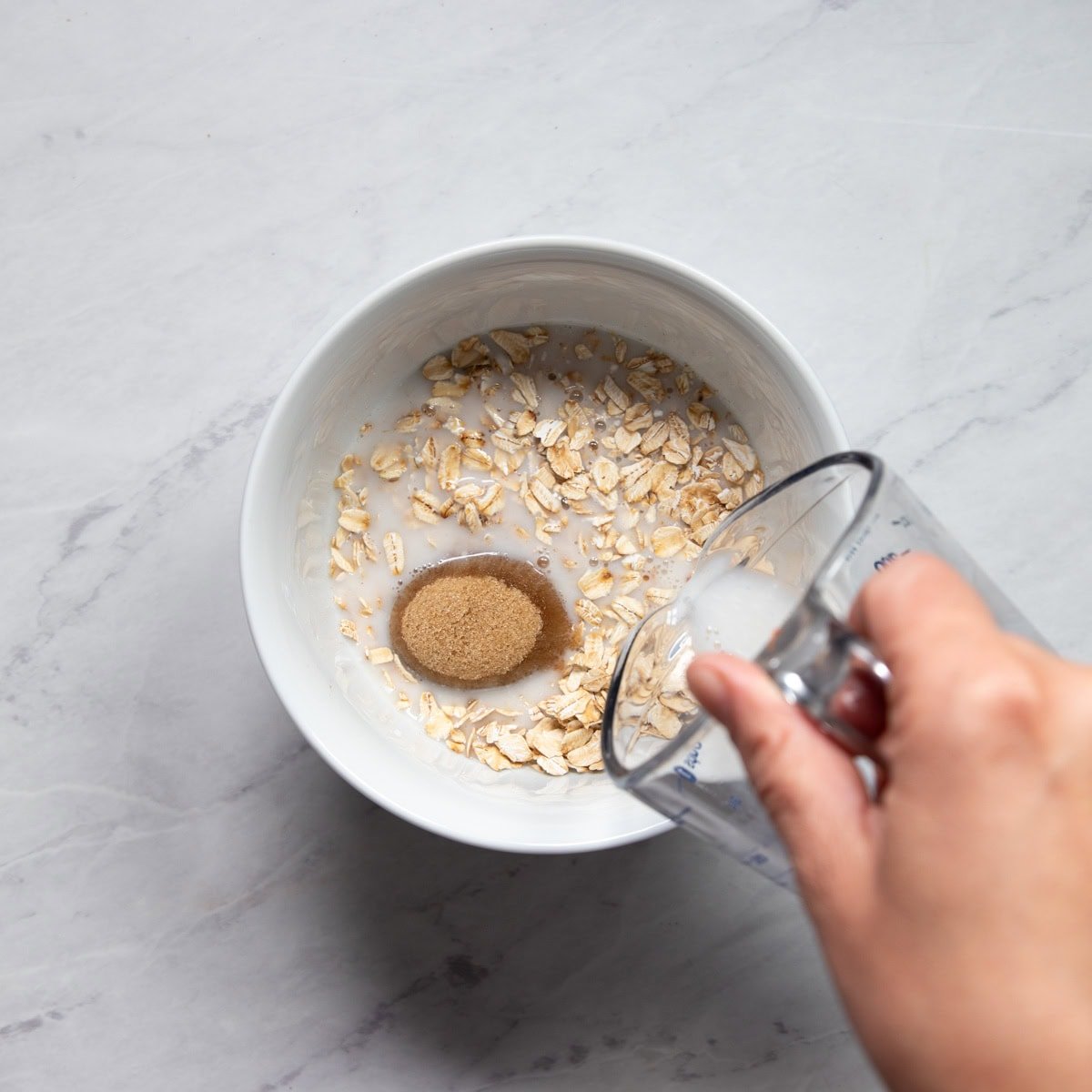 Pouring low FODMAP milk into a bowl containing rolled oats, brown sugar, and vanilla.