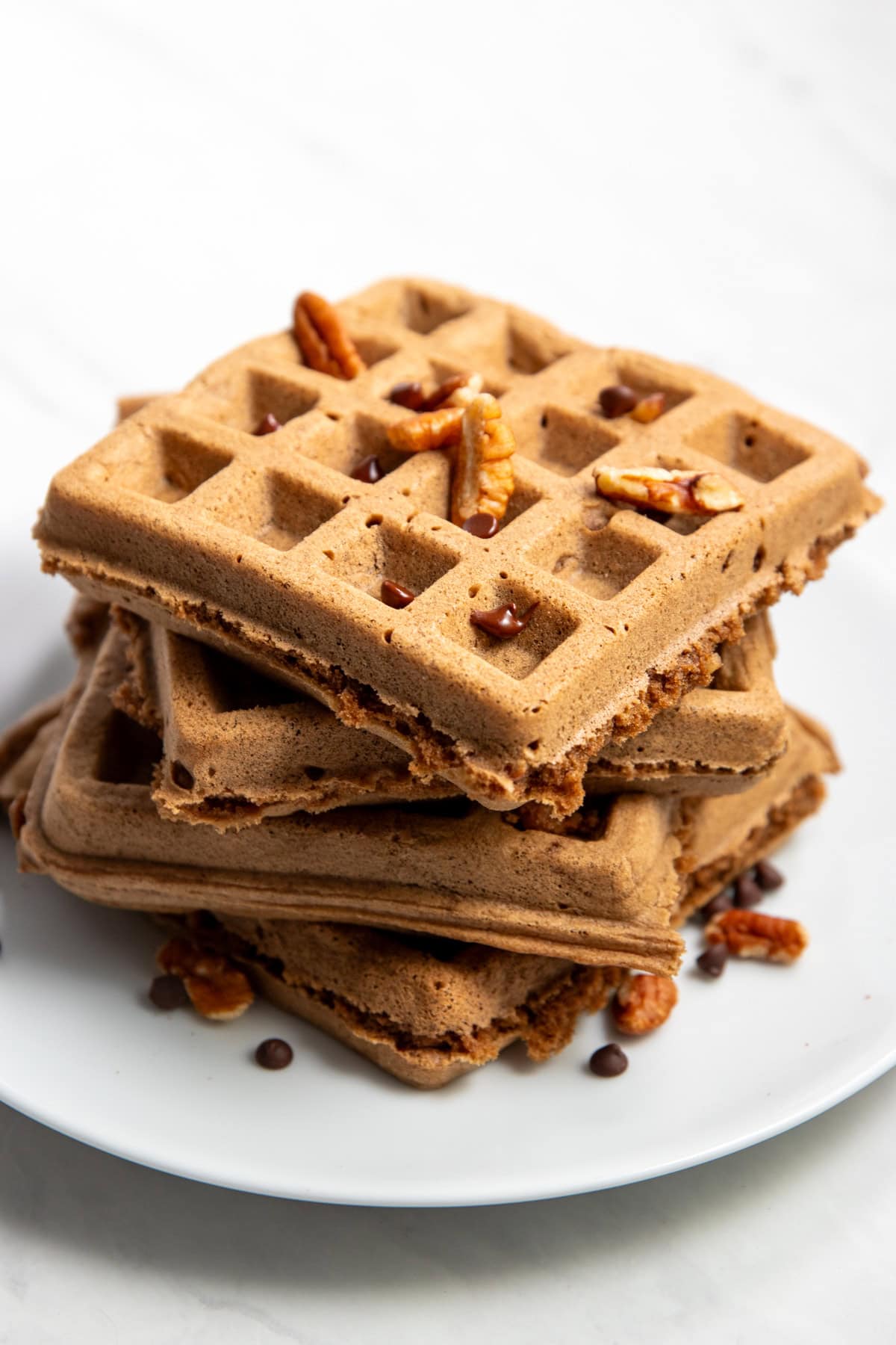 A staggered stack of chocolate waffles on white plate. Chopped pecans and mini chocolate chips are scattered on top of and surrounding the waffles.