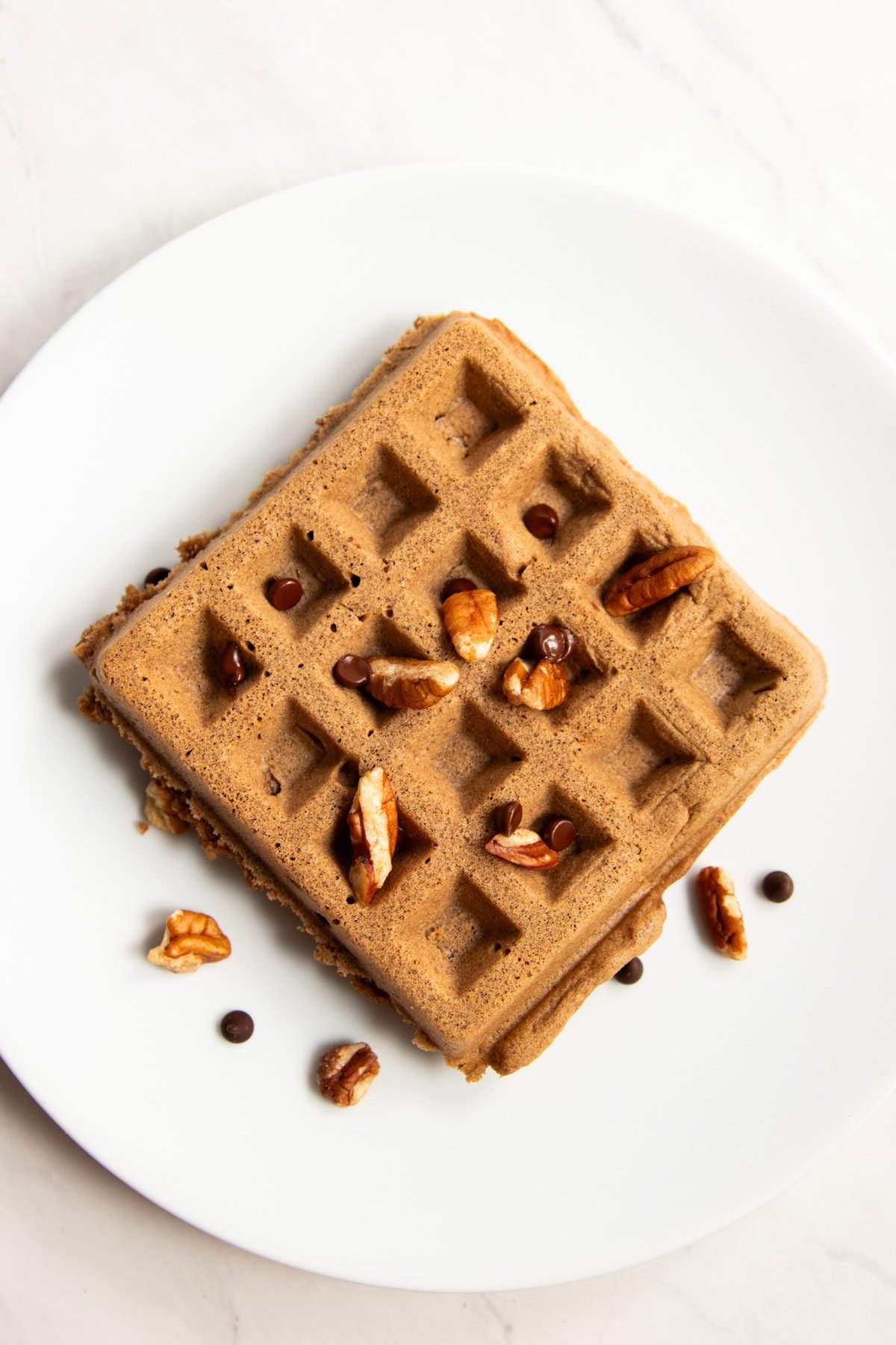 A single chocolate waffle on a plate topped with a few chopped pecans and mini chocolate chips.