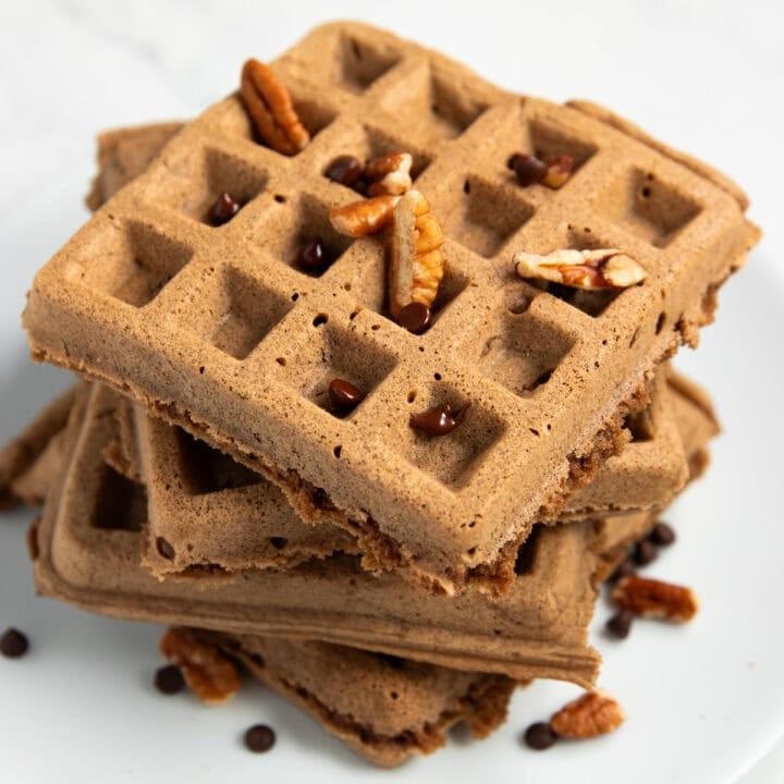 A stack of 4 chocolate pecan waffles staggered on a white plate. There are chopped pecans and mini chocolate chips scattered on top and surrounding the waffles.