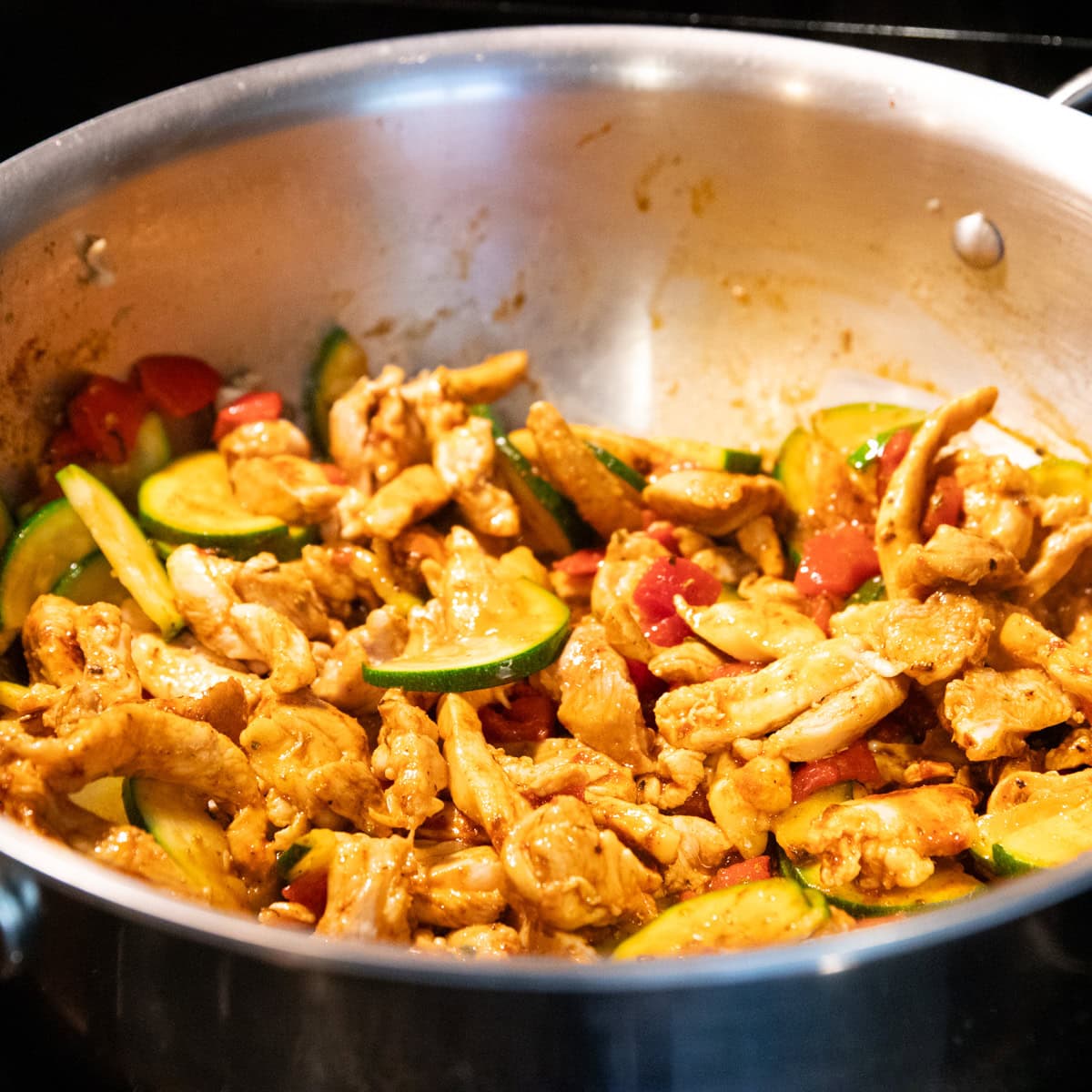 Cooked chicken strips, zucchini, and roasted red pepper are coated with low FODMAP Cajun seasoning in a stainless steel skillet.