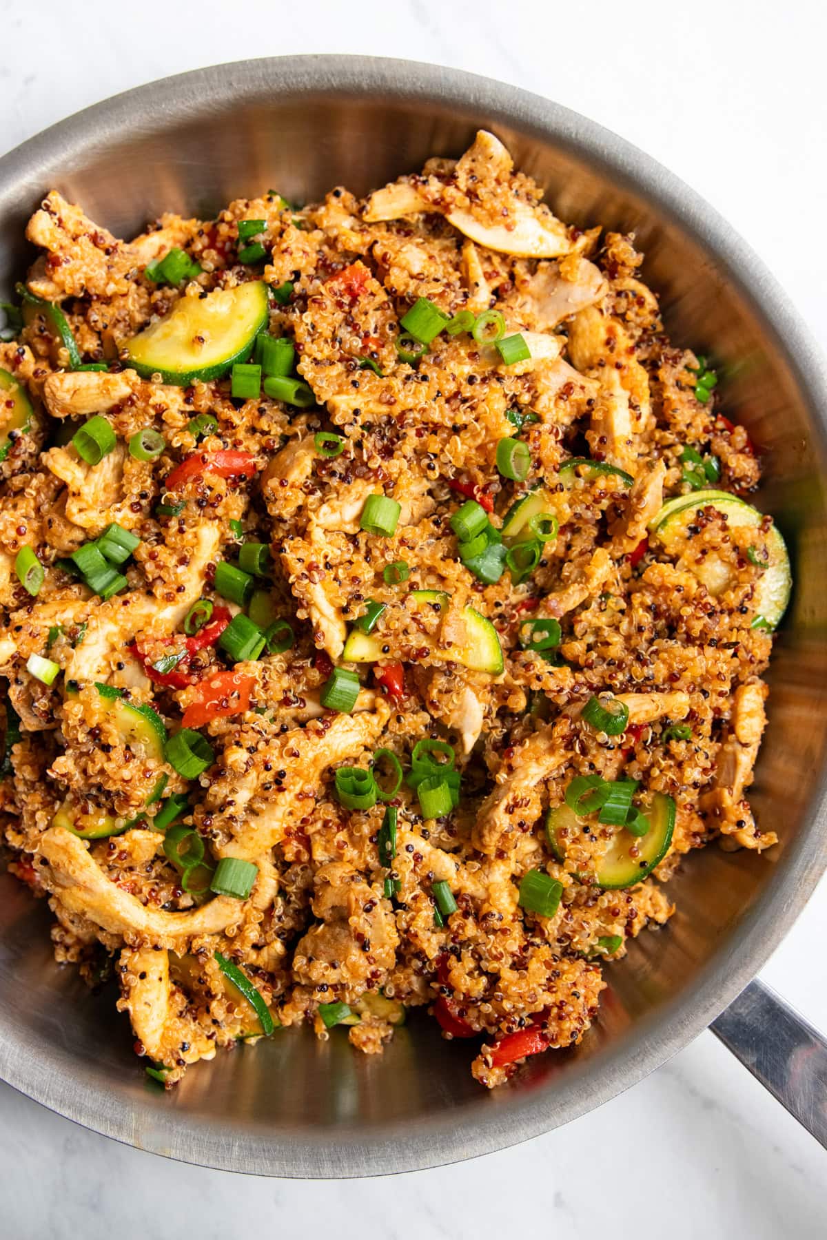 Looking down into a stainless steel skillet filled with strips of chicken, cooked quinoa, sauteed zucchini slices and roasted red peppers.