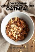 Looking into a bowl of oatmeal sitting on a wood tray next to a tan linen napkin. Over the tan napkin, a black text overlay says "Low FODMAP Chocolate Peanut Butter Oatmeal."