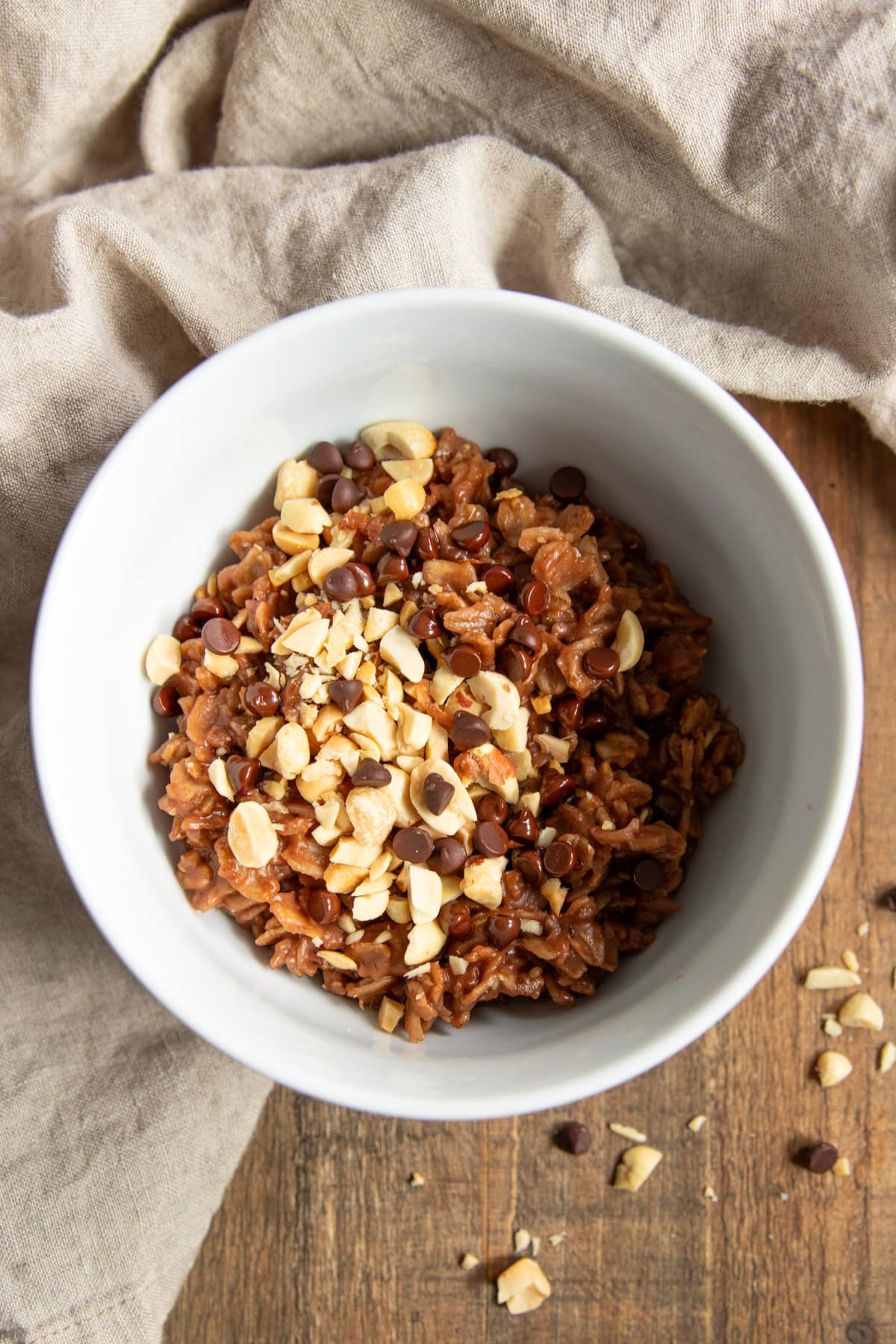 A bowl of chocolatey oatmeal sits on a wood tray. It's surrounded by a tan linen napkin. Chopped peanuts and mini chocolate chips top the oatmeal and are scattered on the wood tray surrounding the bowl.