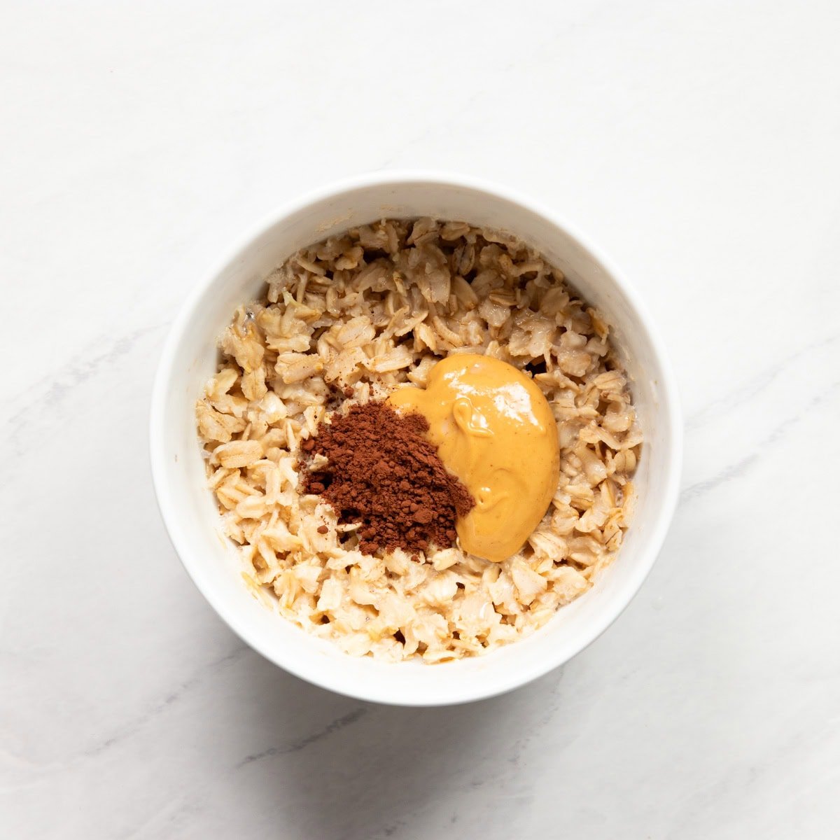 Microwaved rolled oats are topped with peanut butter and cocoa powder, ready for mixing.
