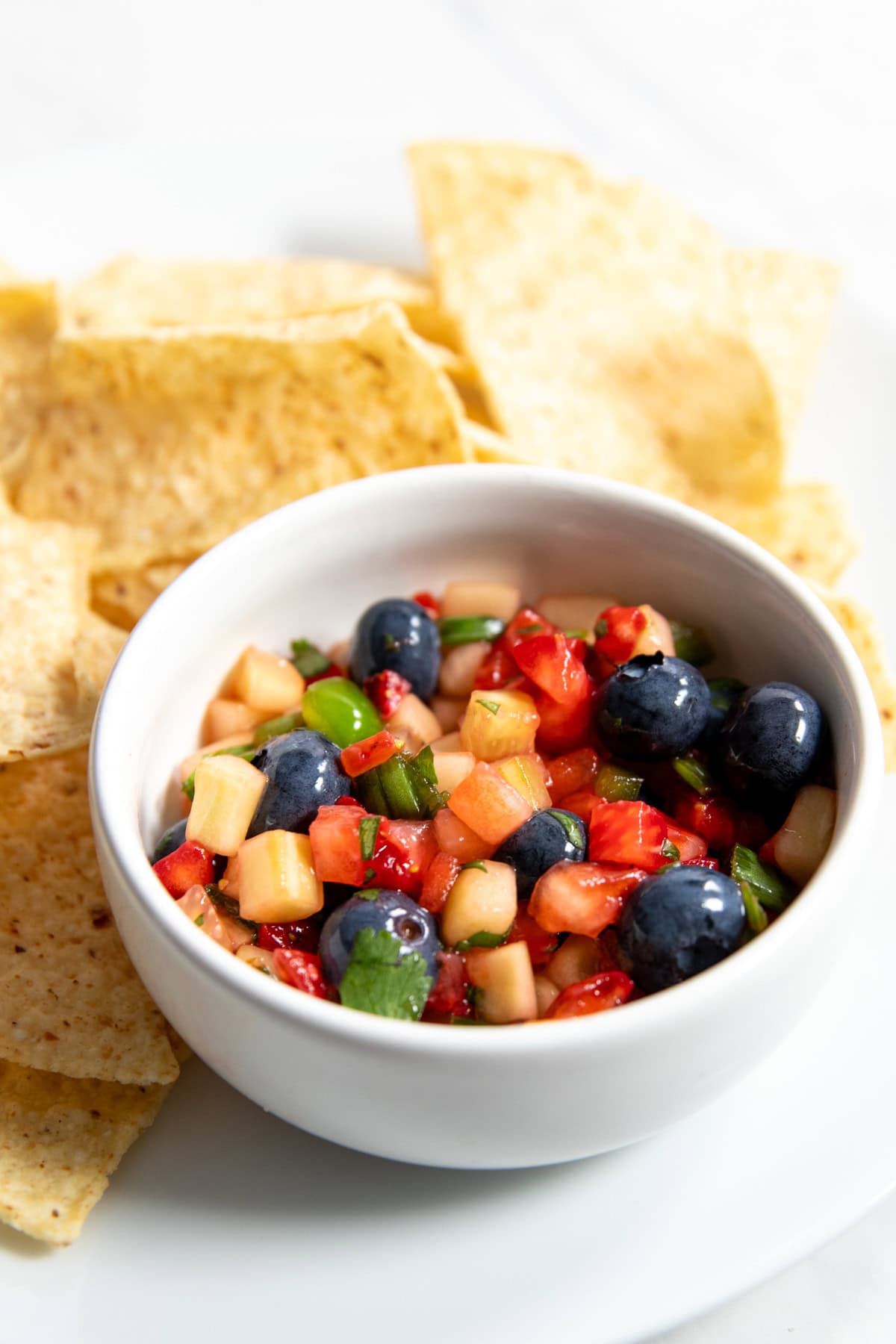 A serving of Low FODMAP Strawberry, Blueberry, and Cucumber Salsa in a small white bowl on a plate surrounded by corn tortilla chips.