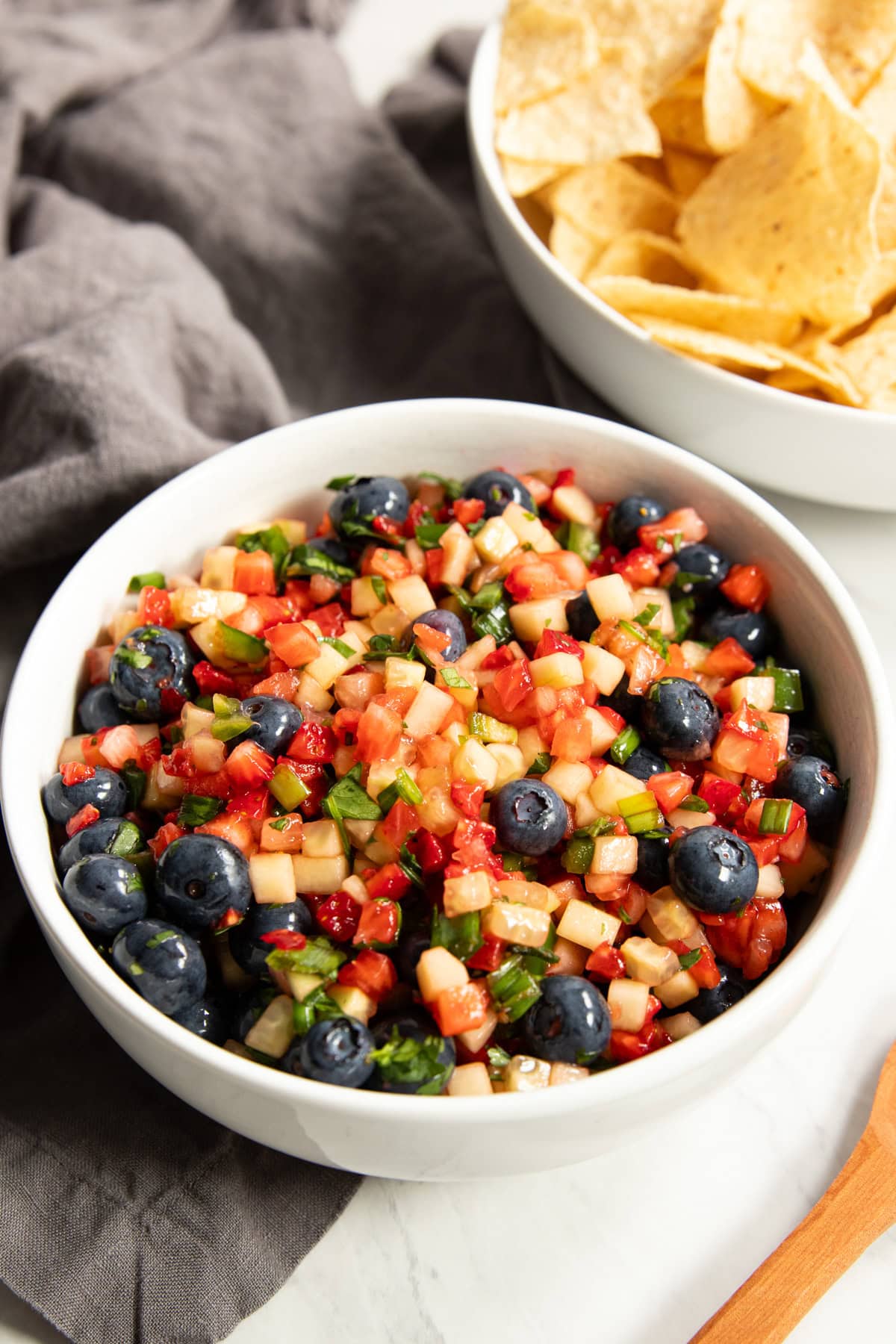A big bowl of fruit salsa made with blueberries, diced strawberries, and diced cucumbers sits on a dark gray linen napkin. There's a wood spoon and a bowl of corn tortilla chips surrounding it. 