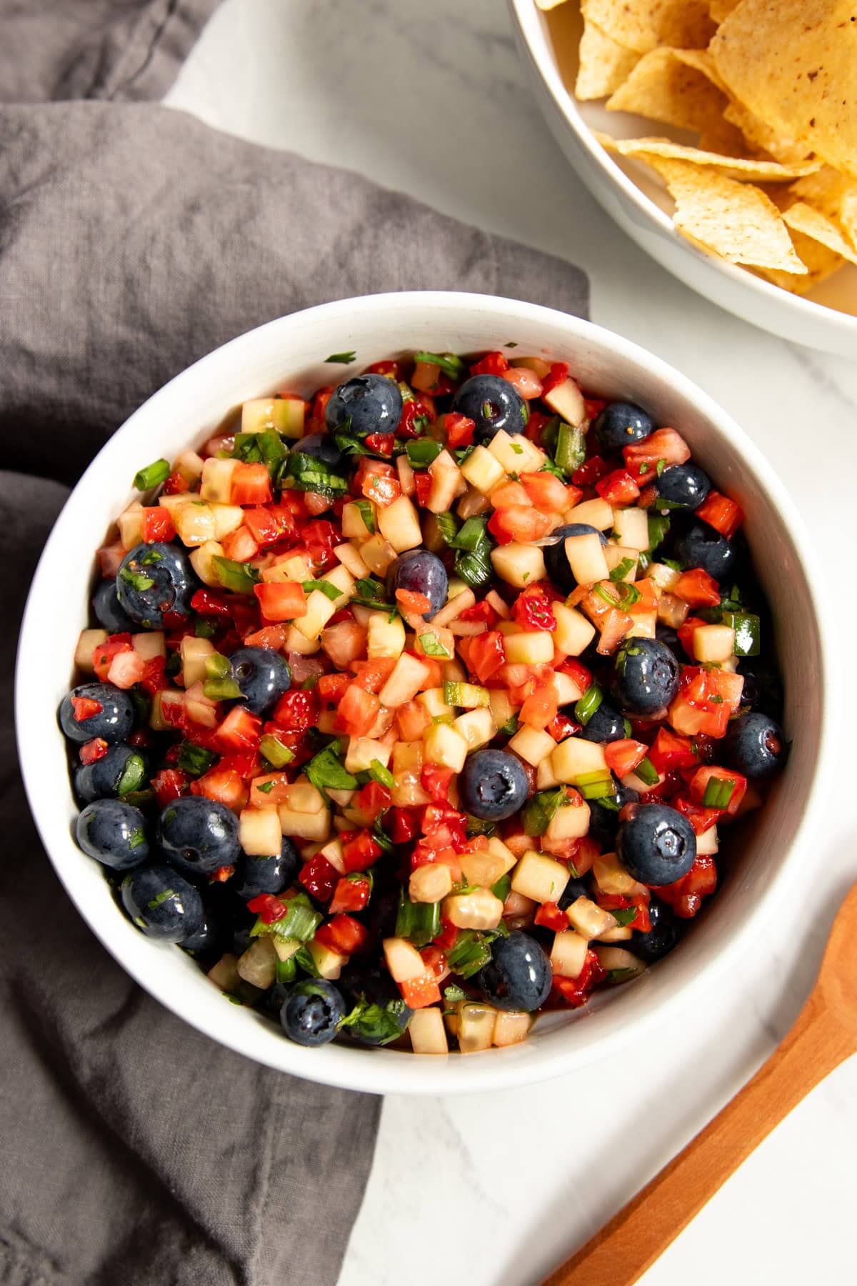 Looking down at a bowl filled with Low FODMAP Strawberry, Blueberry, and Cucumber Salsa sitting on a dark gray linen napkin. There's a bowl of tortilla chips partially in the frame in the right upper corner and a small wooden spoon partially in the frame of the bottom right corner.