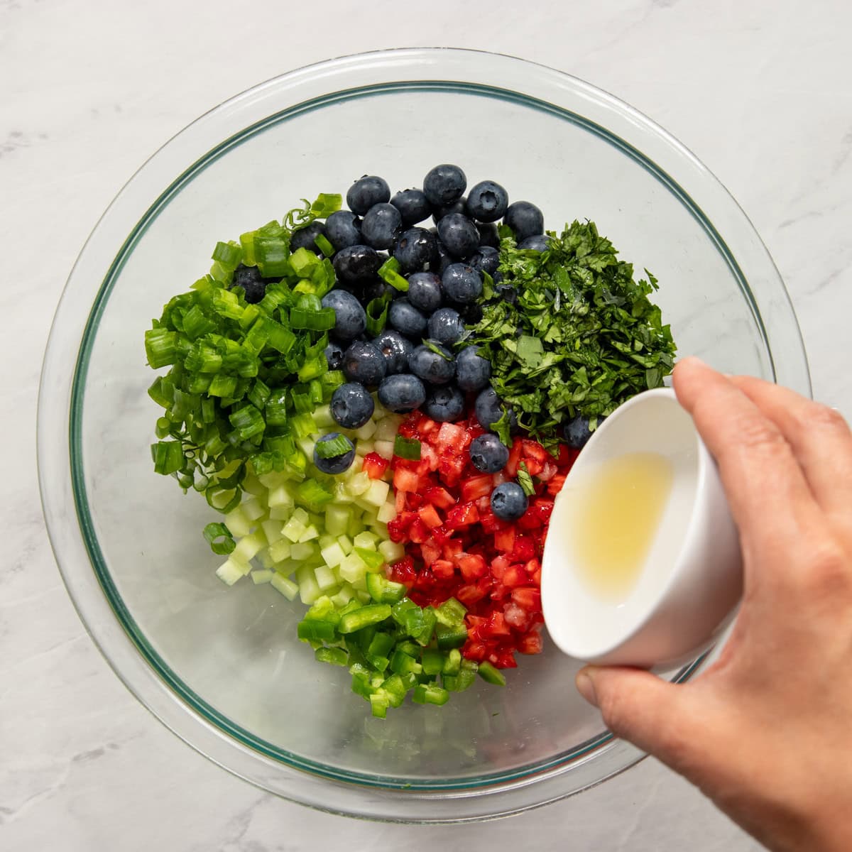 Pouring lime juice into a large glass mixing bowl filled with diced strawberries, diced cucumbers, blueberries, minced jalapeno, chopped cilantro and salt.