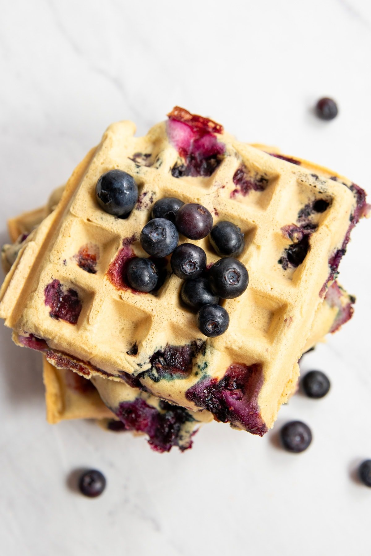A stack of low FODMAP blueberry waffles on a white marble countertop. There are fresh blueberries scattered on top of the waffles, as well as around the base of the waffle tower.