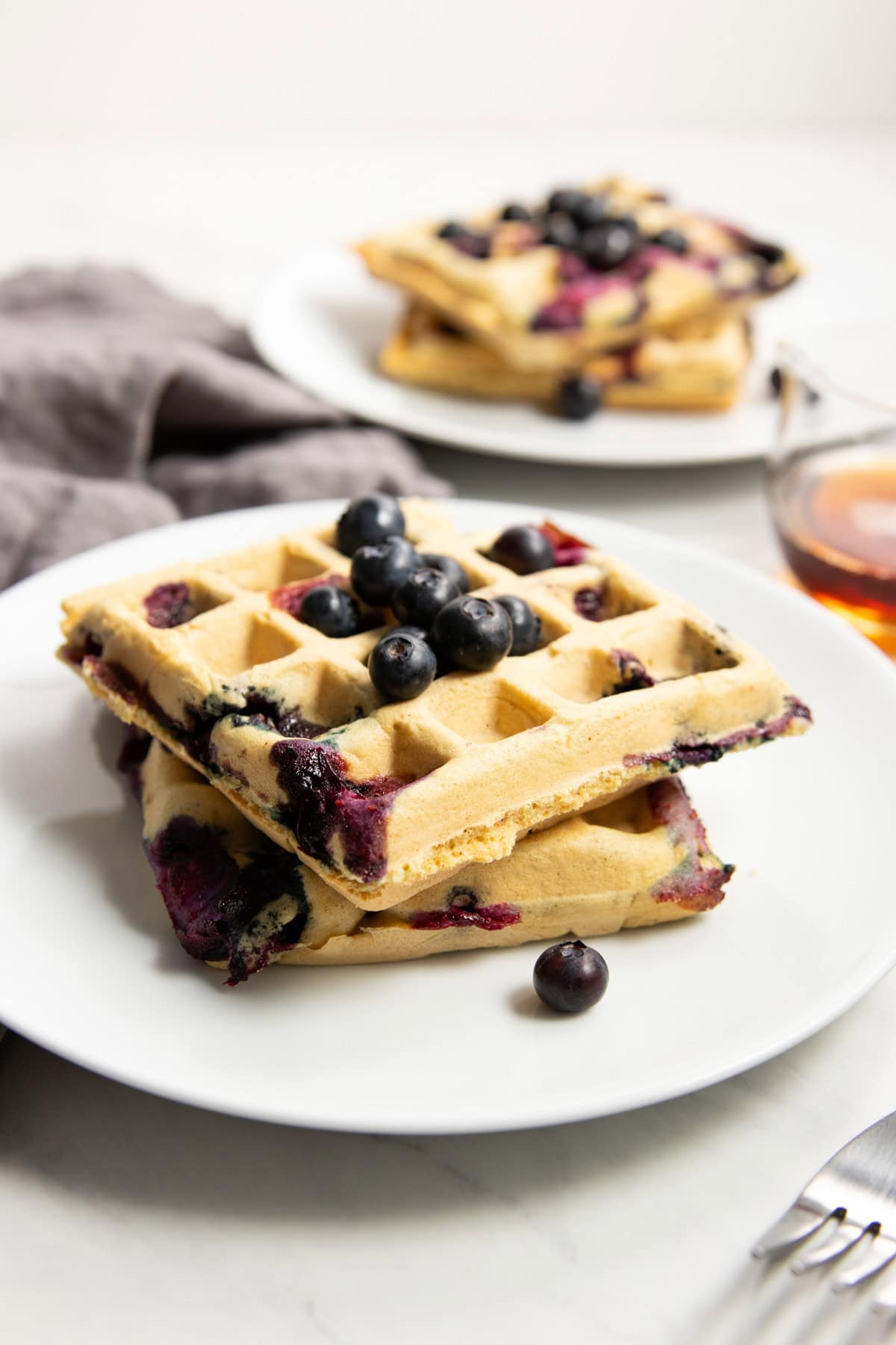 A plate of two blueberry waffles topped with fresh blueberries. There's a gray linen napkin, small cup of maple syrup, and a second plate of waffles staggered artistically in the background.