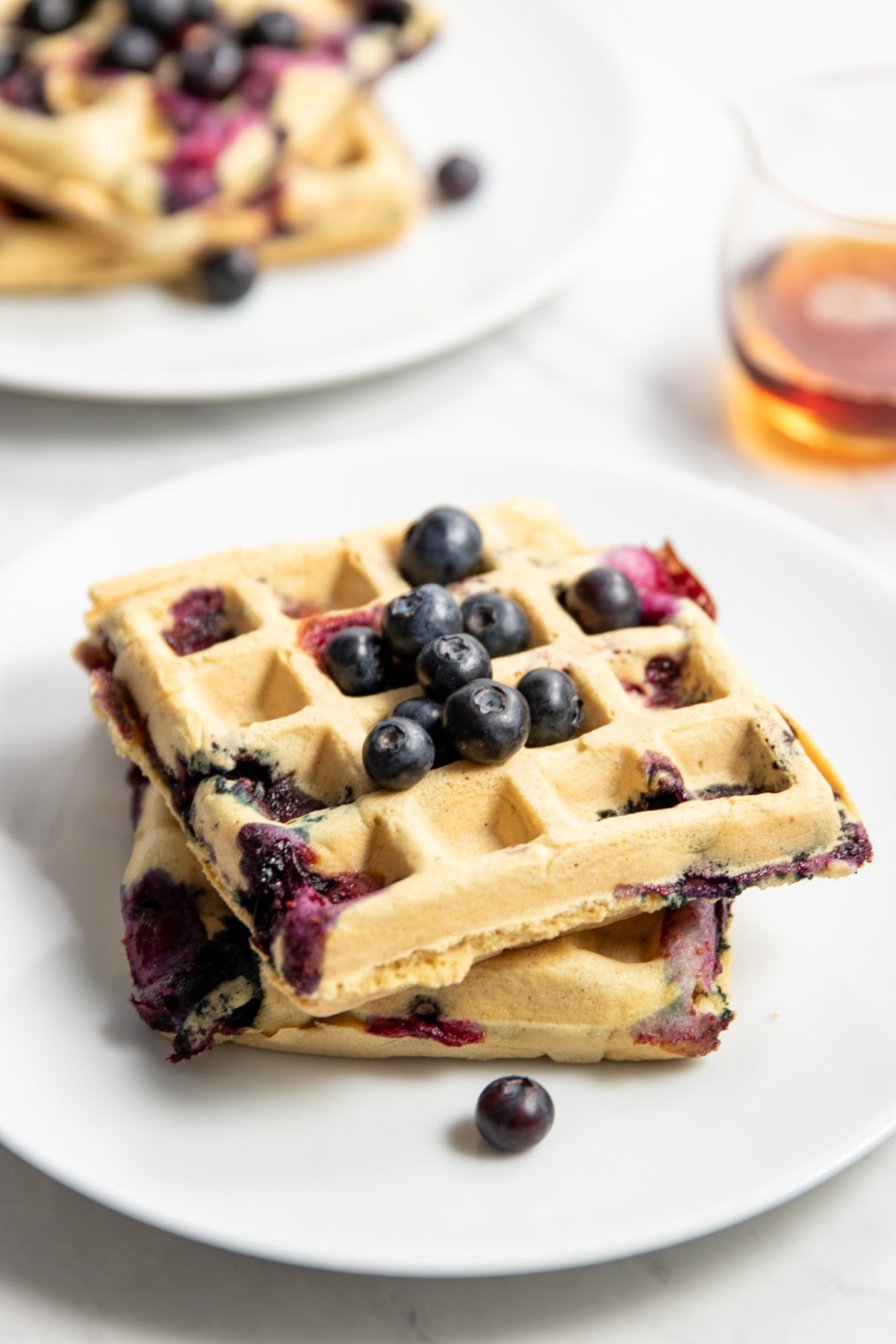 Two plates each with two square blueberry waffles are staggered on a white counter. There's a small glass jar of maple syrup sitting in between the two plates.