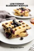 Two plates of blueberry waffles staggered on a white counter with maple syrup and a dark gray linen napkin. In the white space, black text reads "Low FODMAP Blueberry Waffles."