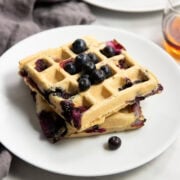 Two square blueberry waffles sitting on a white plate topped with fresh blueberries. A small cup of maple syrup sits off to the right and a dark gray linen napkin surrounds the left side of the plate.