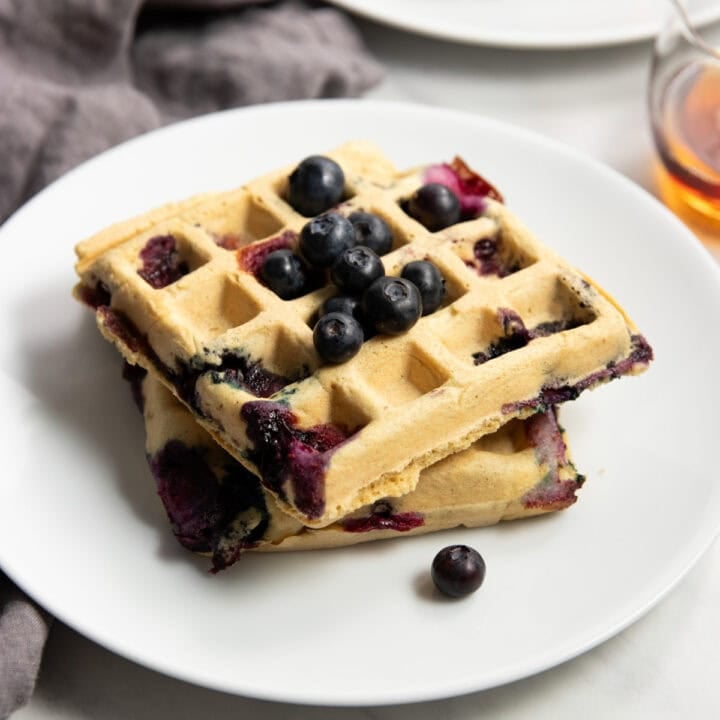 Two square blueberry waffles sitting on a white plate topped with fresh blueberries. A small cup of maple syrup sits off to the right and a dark gray linen napkin surrounds the left side of the plate.
