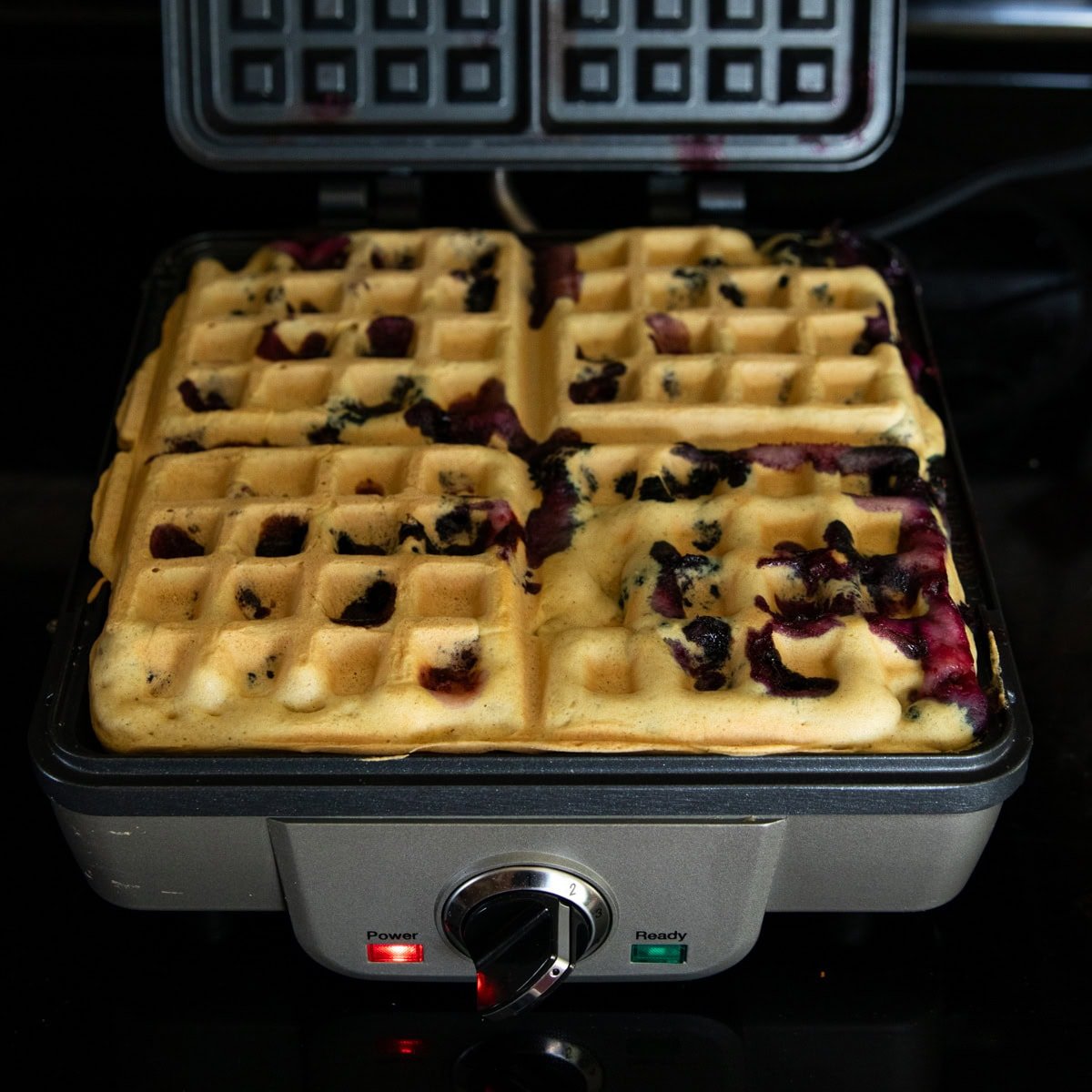 An almost cooked blueberry waffle sitting on an open waffle iron.