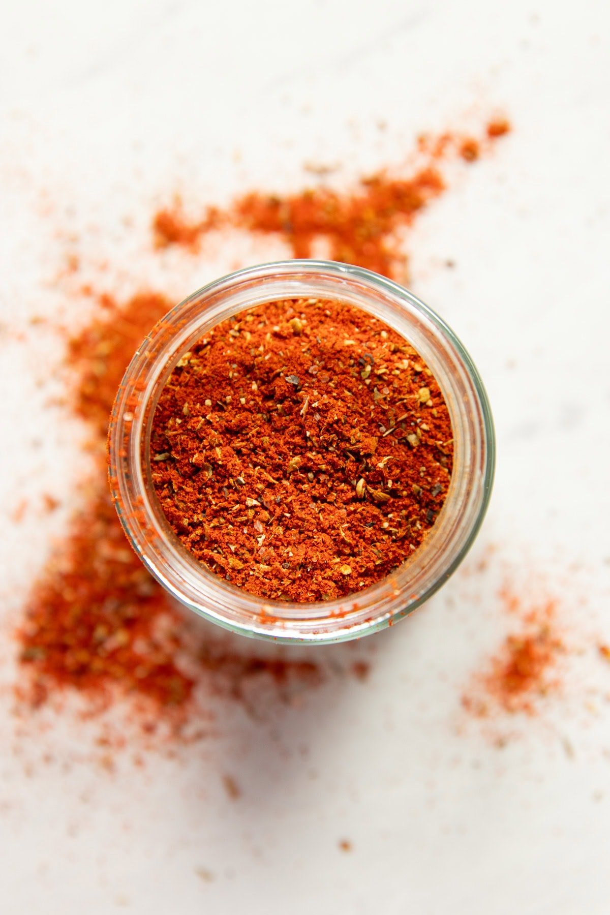 Looking down onto a small jar filled with a red spice blend. Paprika and other herbs and spices are spilled around the jar.