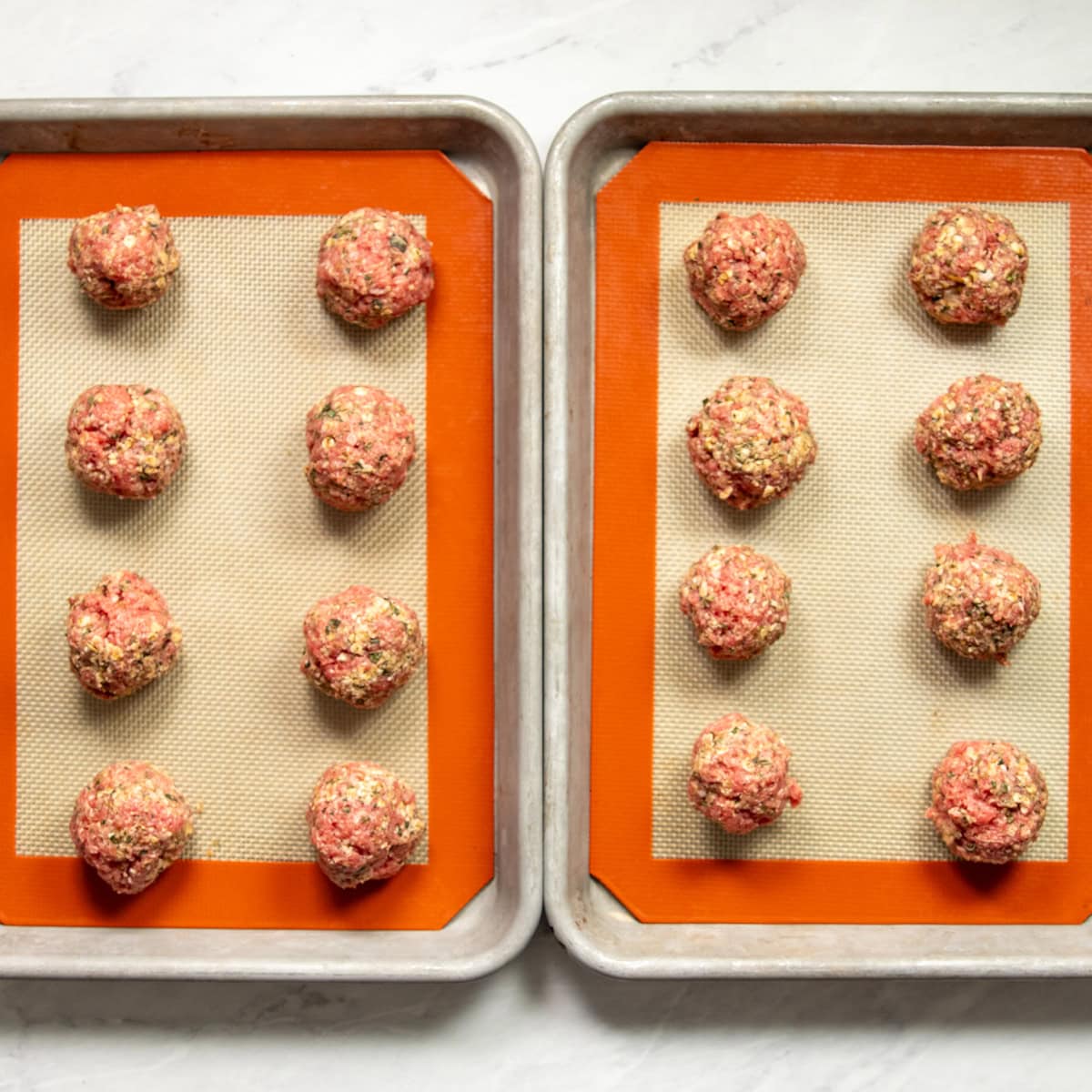 Sixteen meatballs are rolled out and ready to bake on two side by side baking sheets lined with silicone baking mats. 