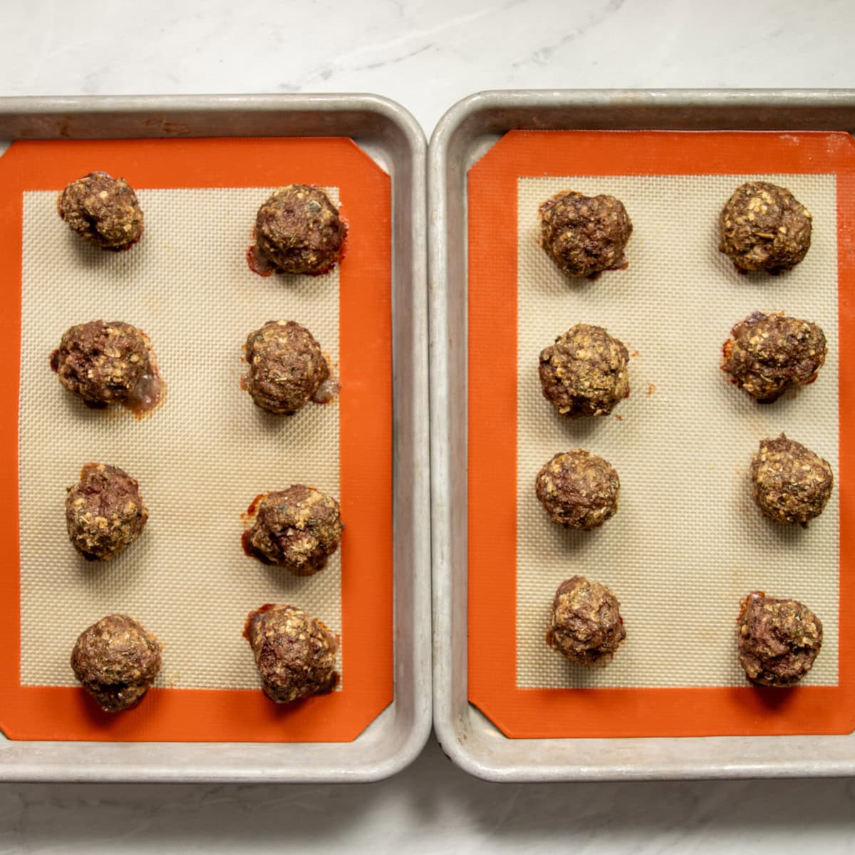 Two baking sheets lined with silicone mats have four rows of four baked Italian beef meatballs.