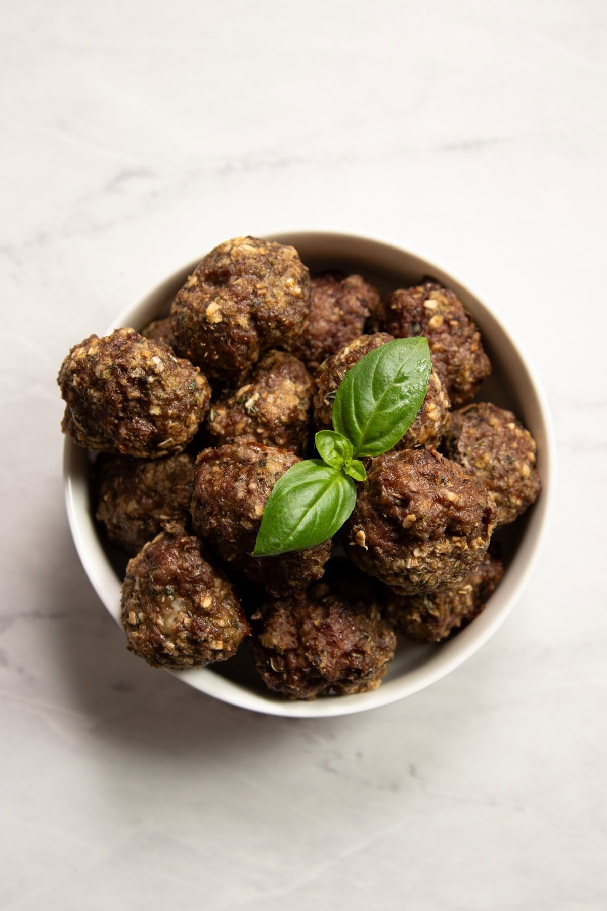 Looking down into a white bowl filled with Low FODMAP Italian Beef Meatballs sitting on a white marble counter. A basil leaf garnish artistically sits on top of the meatballs. 