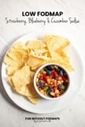 Looking down at a plate filled with corn tortilla chips and a small bowl of fruit salsa. In the white space above, black text reads "Low FODMAP Strawberry, Blueberry, and Cucumber Salsa"