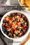 Looking down at a bowl of fruit salsa surrounded by a gray linen napkin, a small wood spoon, and a bowl of tortilla chips. A black text overlay reads "Low FODMAP Strawberry, Blueberry, and Cucumber Salsa."