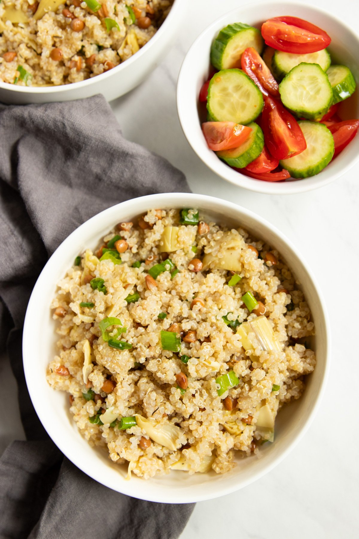 Looking over three bowls scattered on a white counter with gray linen napkin underneath. Two of the bowls contain lemon quinoa salad with lentils and artichokes and one contains sliced cucumbers and cherry tomatoes. 