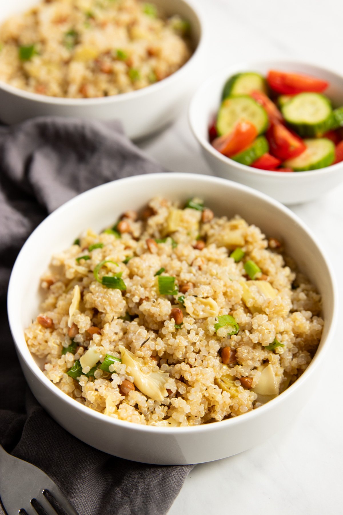 A close up of a bowl filled with Low FODMAP Lemon Quinoa Salad with Lentils and Artichokes sitting on a gray linen napkin. Blurred in the background is a small dish of cherry tomatoes and cucumber slices, as well as second bowl of salad.