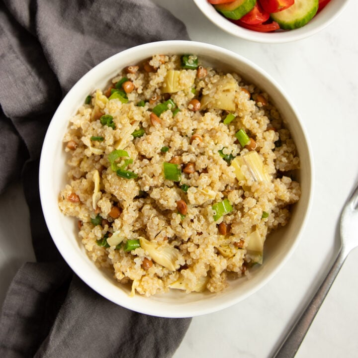A bowl of quinoa salad dotted with chopped artichokes, canned lentils and sliced green onion tops. A small bowl of cucumber slices and cherry tomatoes is off to the side as is a gray linen napkin and fork.