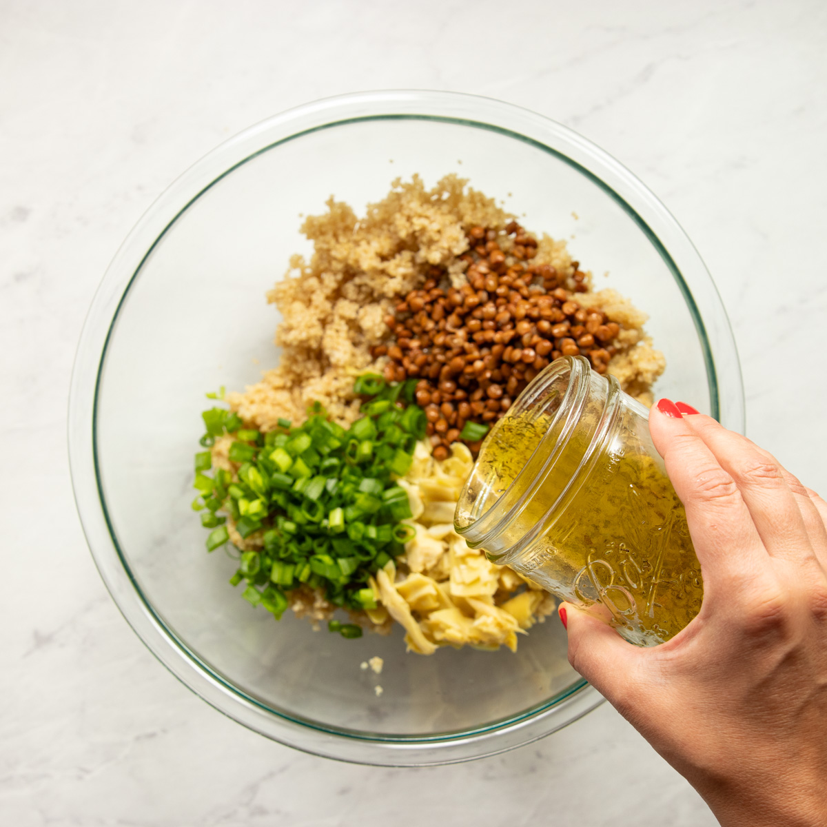 Pouring lemon-thyme dressing over a mixing bowl containing cooked quinoa, canned lentils, canned artichoke hearts, and sliced green onion tops.